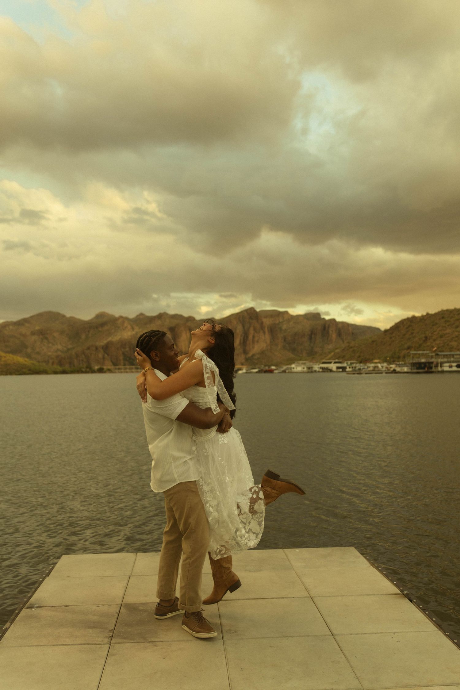 Couple sharing a romantic lift and kiss on a lakeside dock with mountain backdrop at golden sunset.