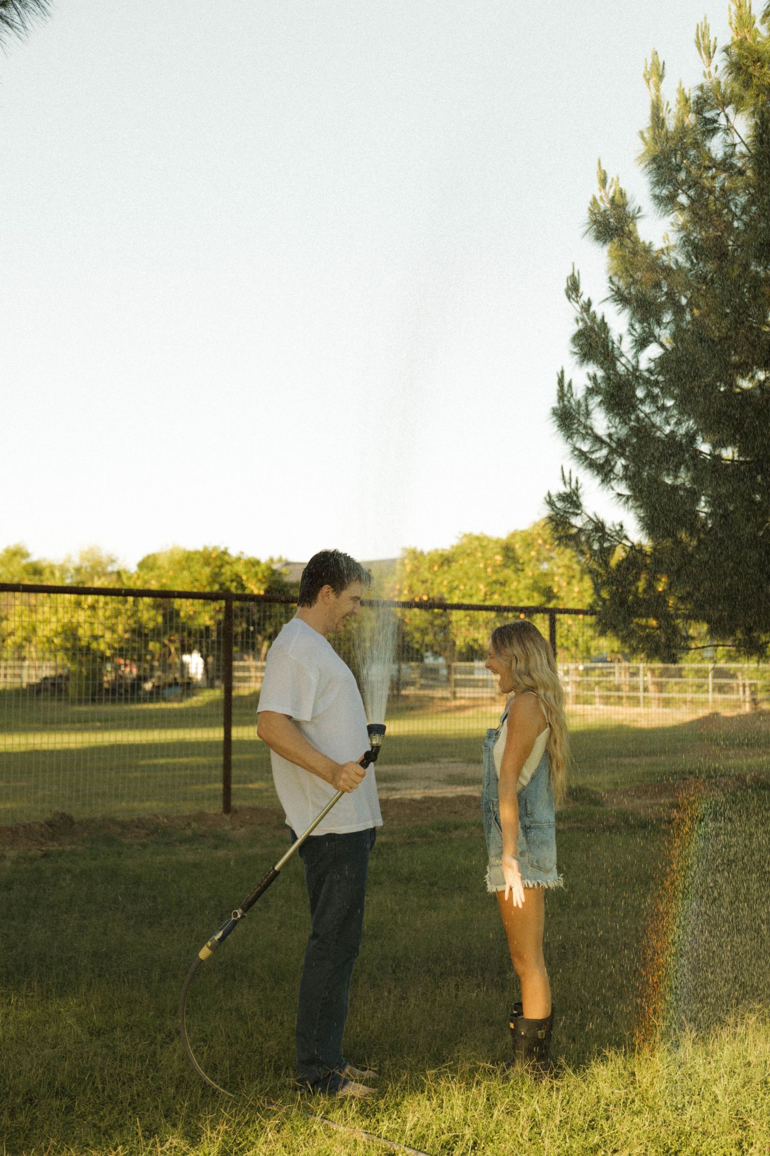 Young couple standing face to face in a sunny backyard while man holds a spraying garden hose at golden hour.