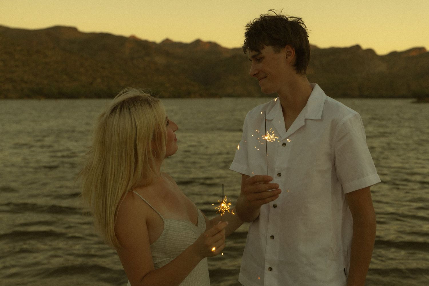 Couple holding sparklers by a lake at sunset with mountains in the background.