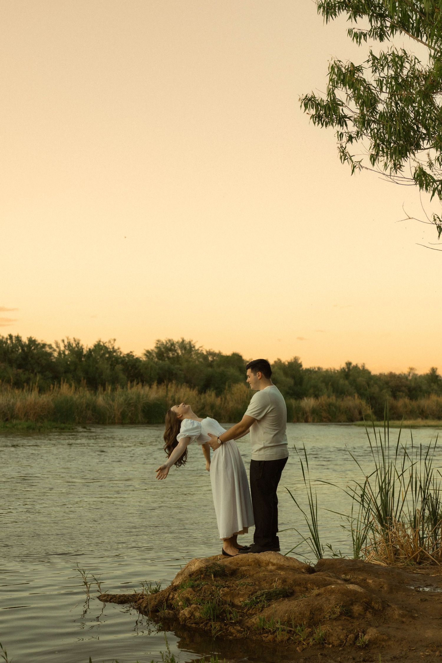 Couple dancing romantically on rocks by a tranquil river at golden sunset, surrounded by reeds and trees.