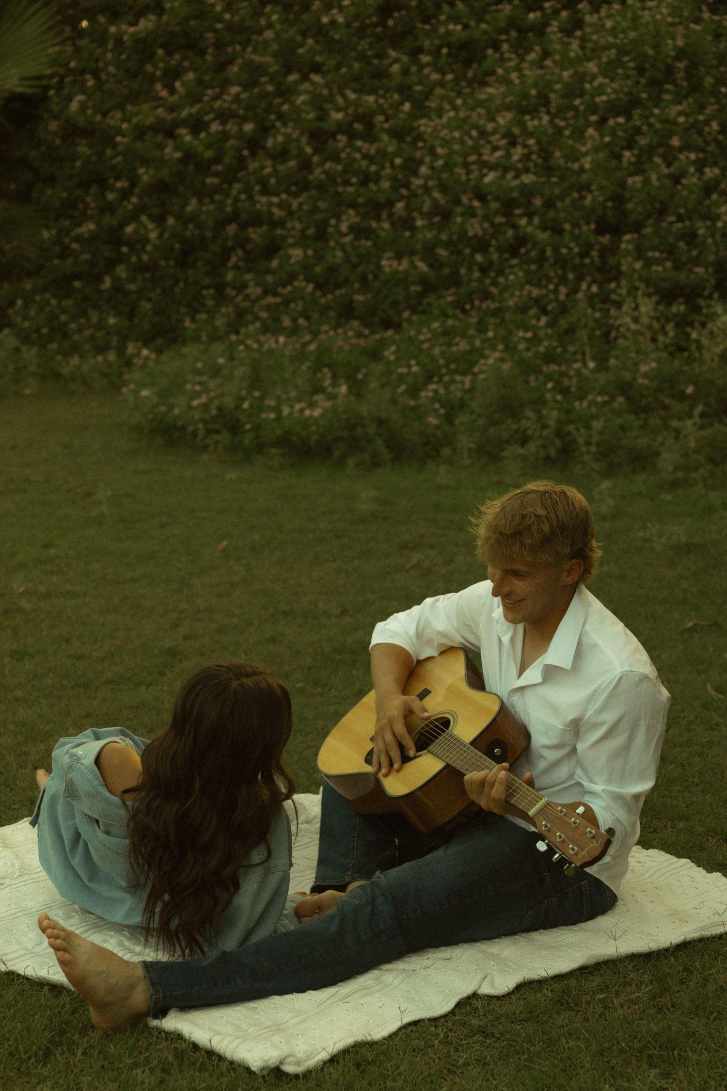 A young couple enjoys a romantic outdoor picnic on a blanket, with a man playing acoustic guitar for a woman on green grass.
