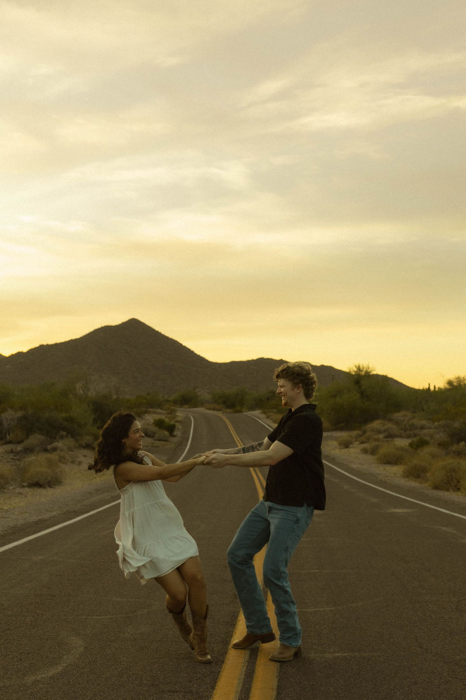 Romantic couple holding hands and spinning on empty desert highway at golden sunset with mountains.