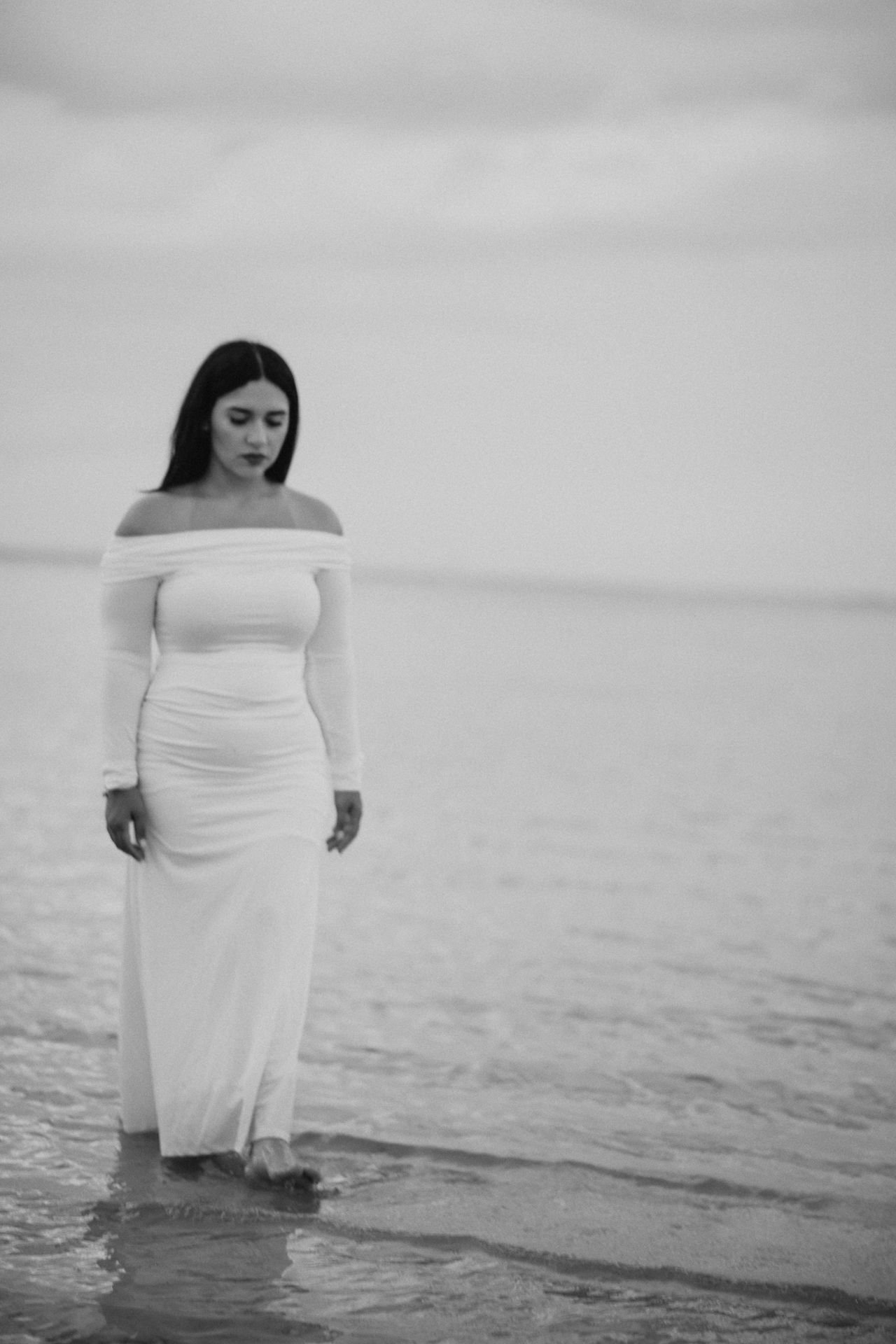 A woman in a fitted off-shoulder white dress stands at the water's edge in a dreamy black and white photo.