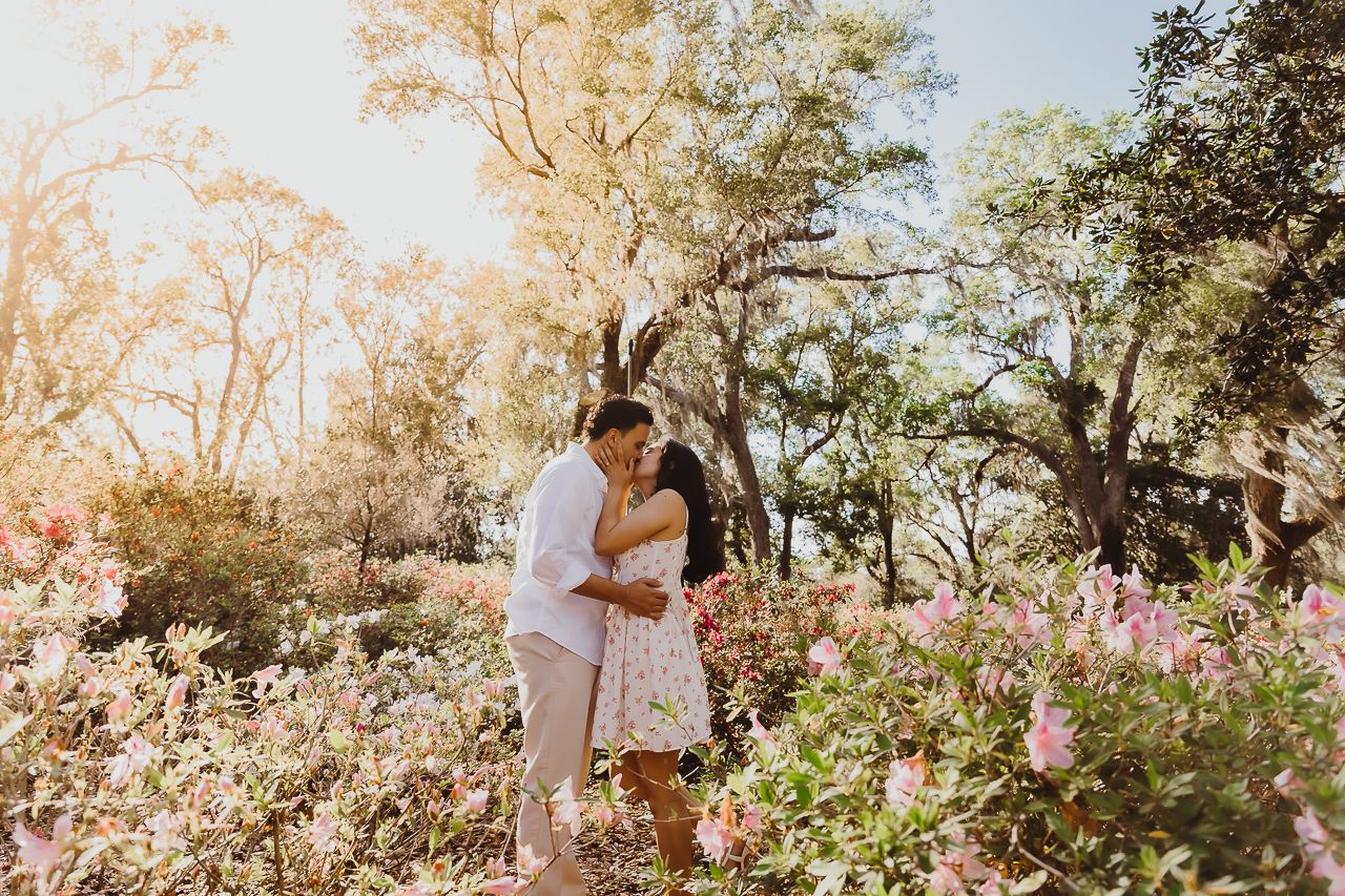 A couple embraces in a sunlit garden filled with blooming pink flowers and towering trees during golden hour.