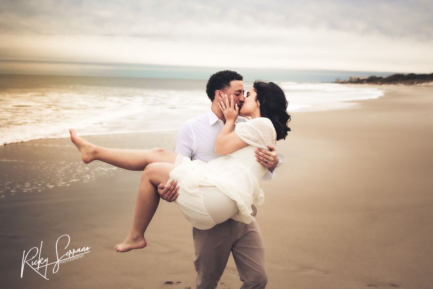 Romantic couple embraces on a misty beach at sunset, with waves and coastline visible in the background.