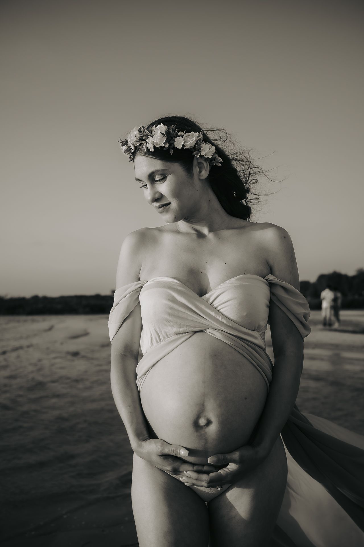 A black and white maternity photo shows an expectant mother in a floral crown and white dress at the beach.