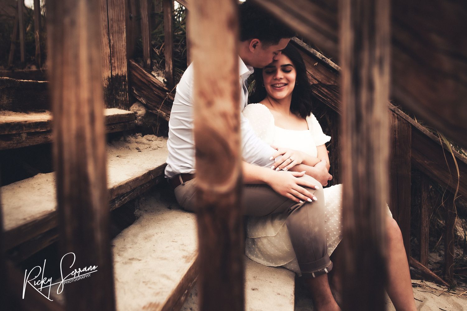 A maternity photo taken through wooden posts shows an expectant mother in a white dress sitting on rustic steps.