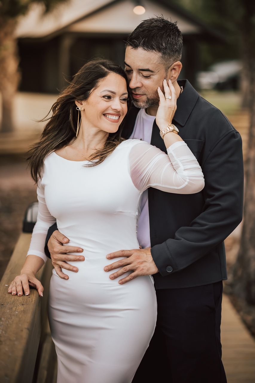 A romantic couple embraces outdoors, with woman in white dress and man in dark suit sharing an intimate moment.