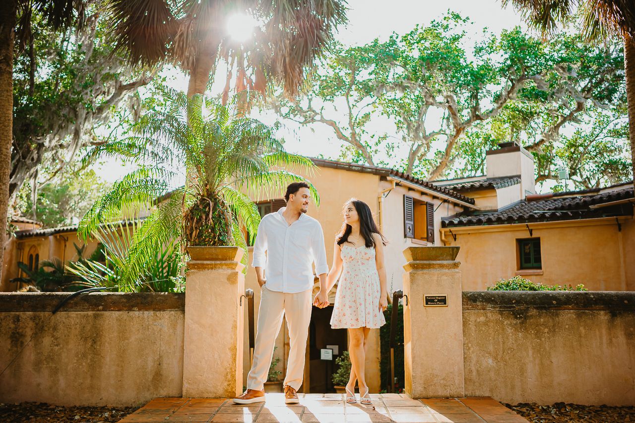 A couple stands together in a sunlit Mediterranean-style courtyard surrounded by palm trees and stucco walls.