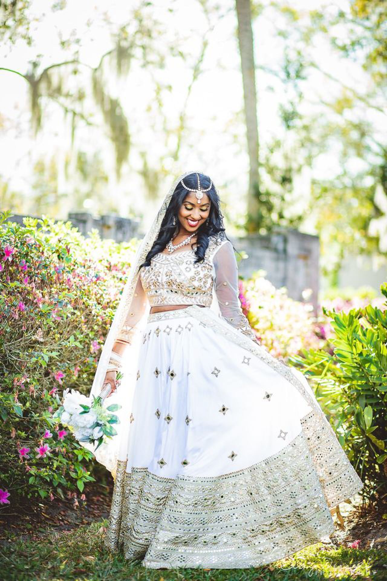 Bride in ornate white and gold traditional Indian lehenga twirling in garden setting.