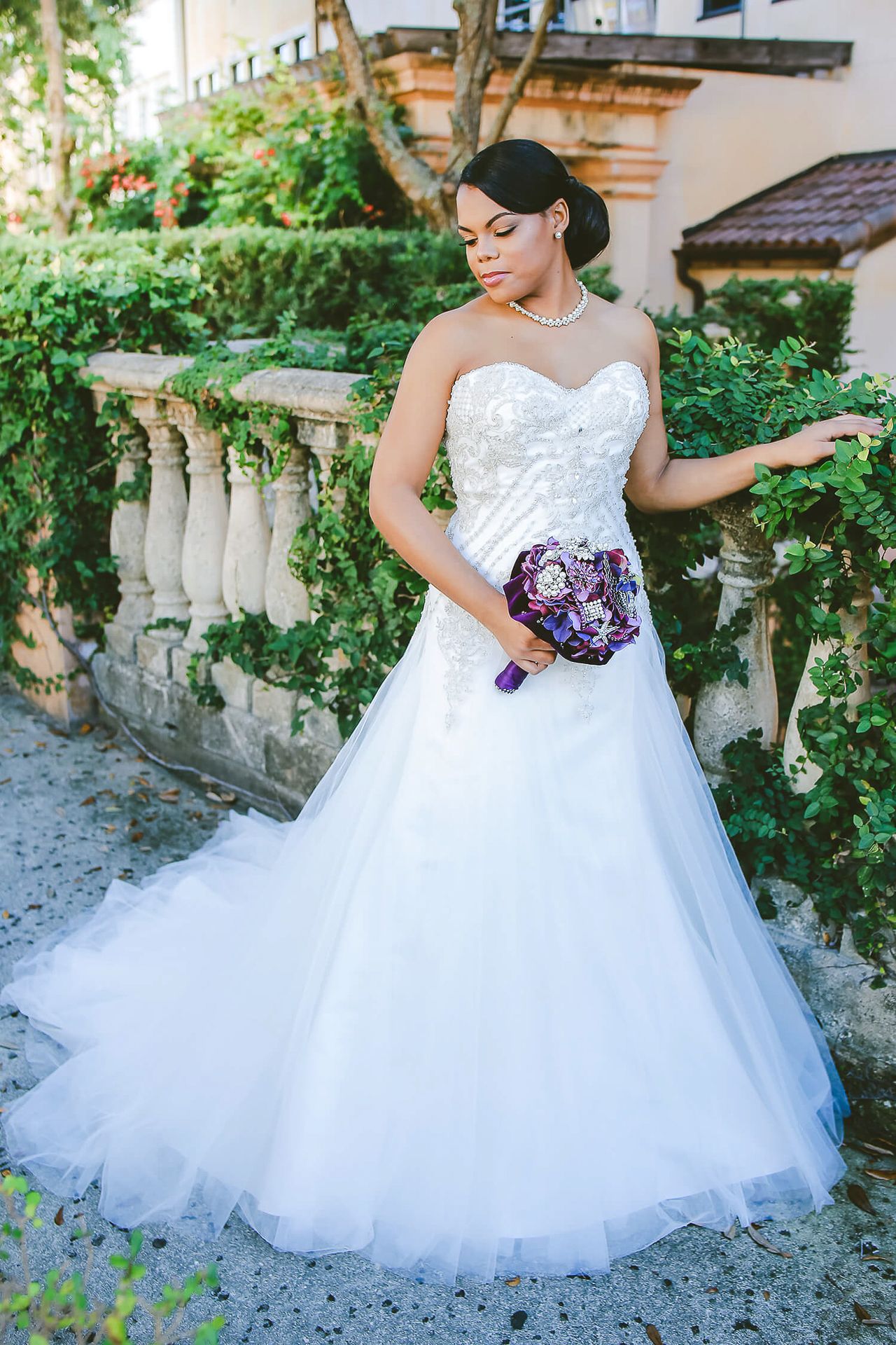 A bride in a white strapless ball gown poses outdoors next to stone balustrades with purple flowers in her hand.