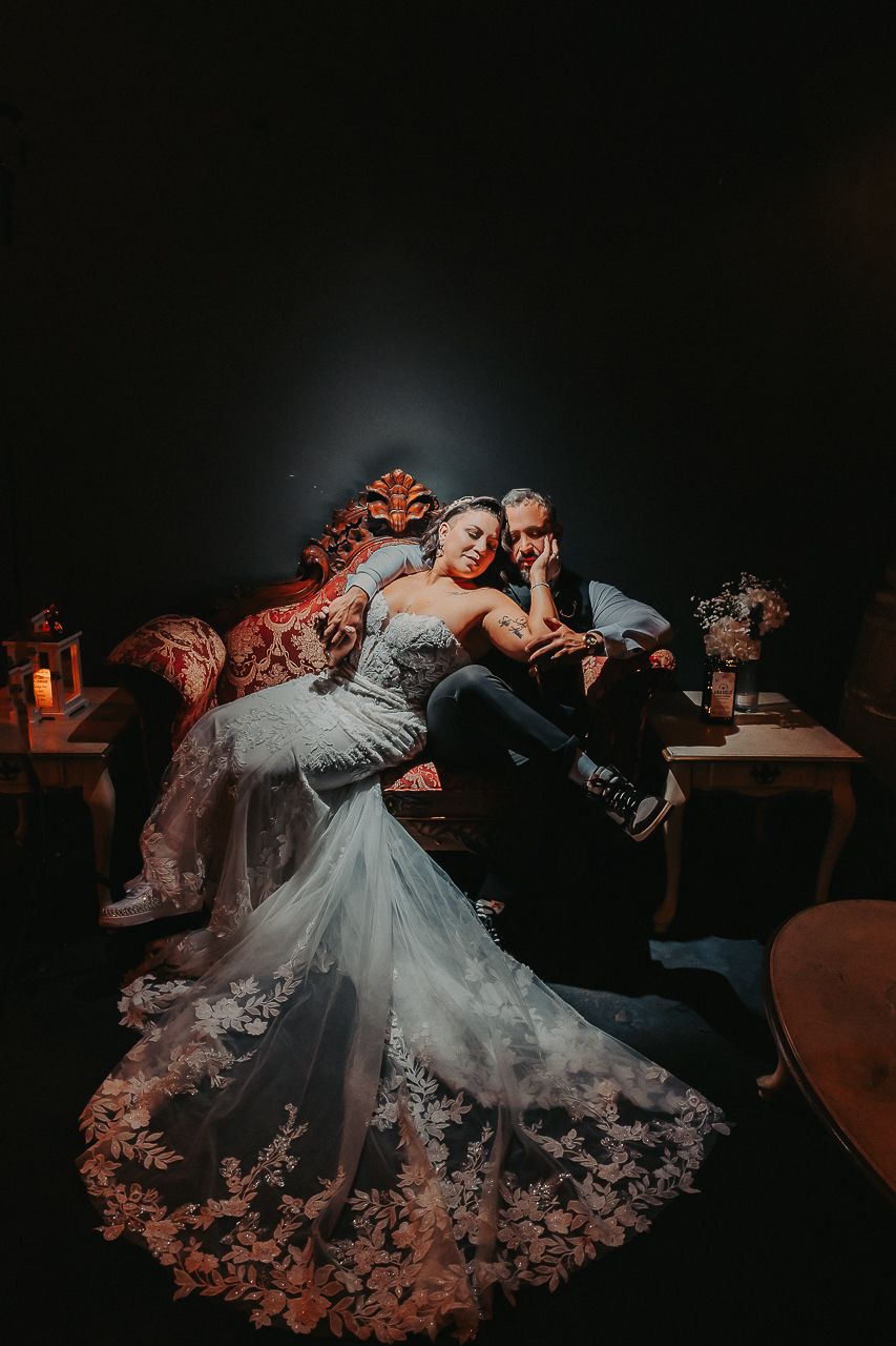A couple relaxes together on a dark sofa, with a stunning lace wedding gown spread beautifully across the frame.