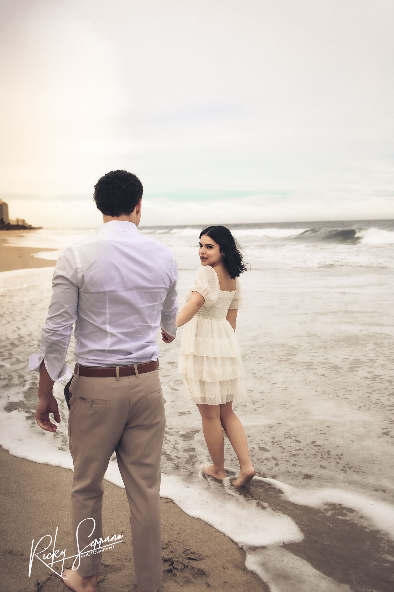A couple shares a romantic moment on a beach with crashing waves and cloudy skies during sunset.