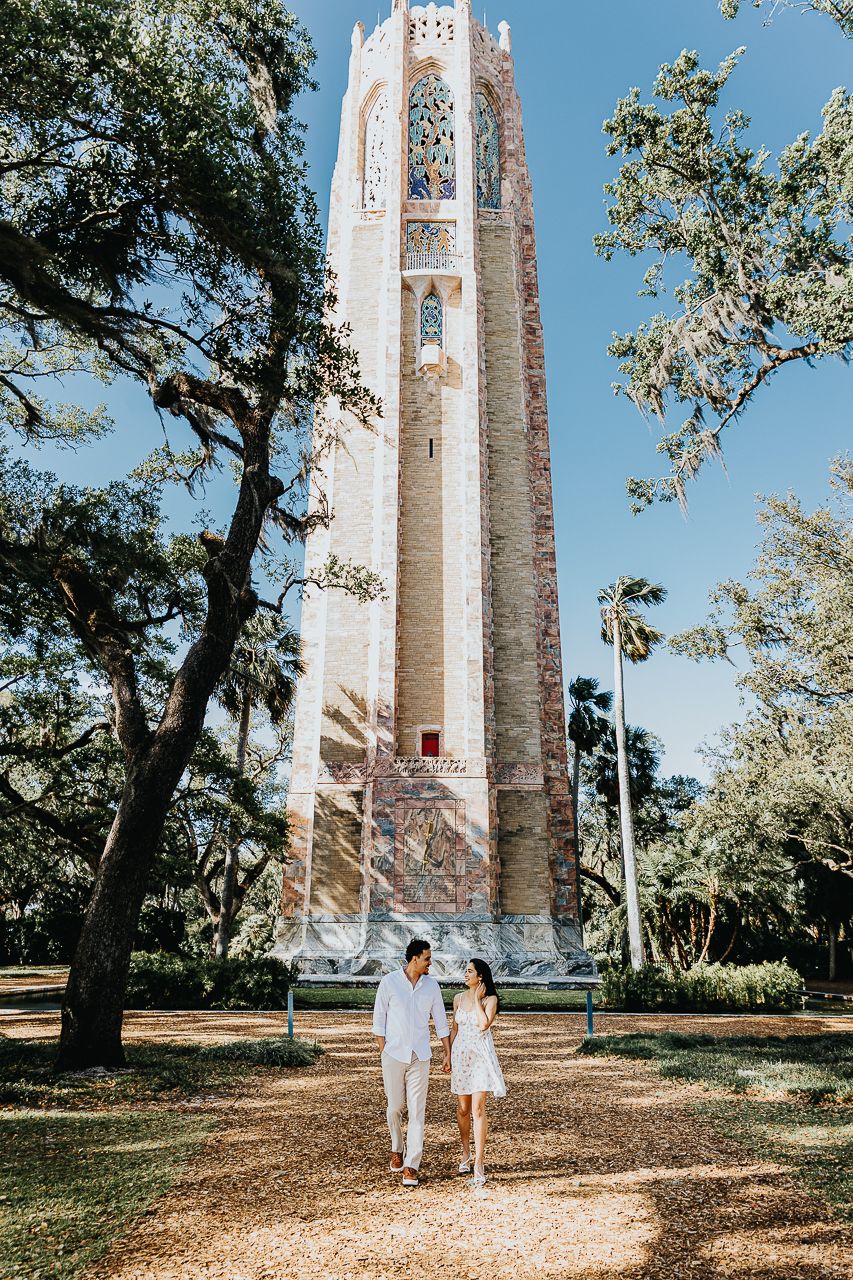 A couple strolls on a path beneath the towering Bok Tower, a historic bell tower surrounded by trees in Florida.
