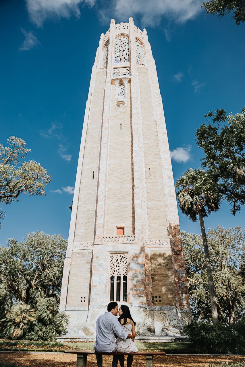 Couple sharing a romantic moment while sitting on a bench in front of a tall white Gothic bell tower on a sunny day.