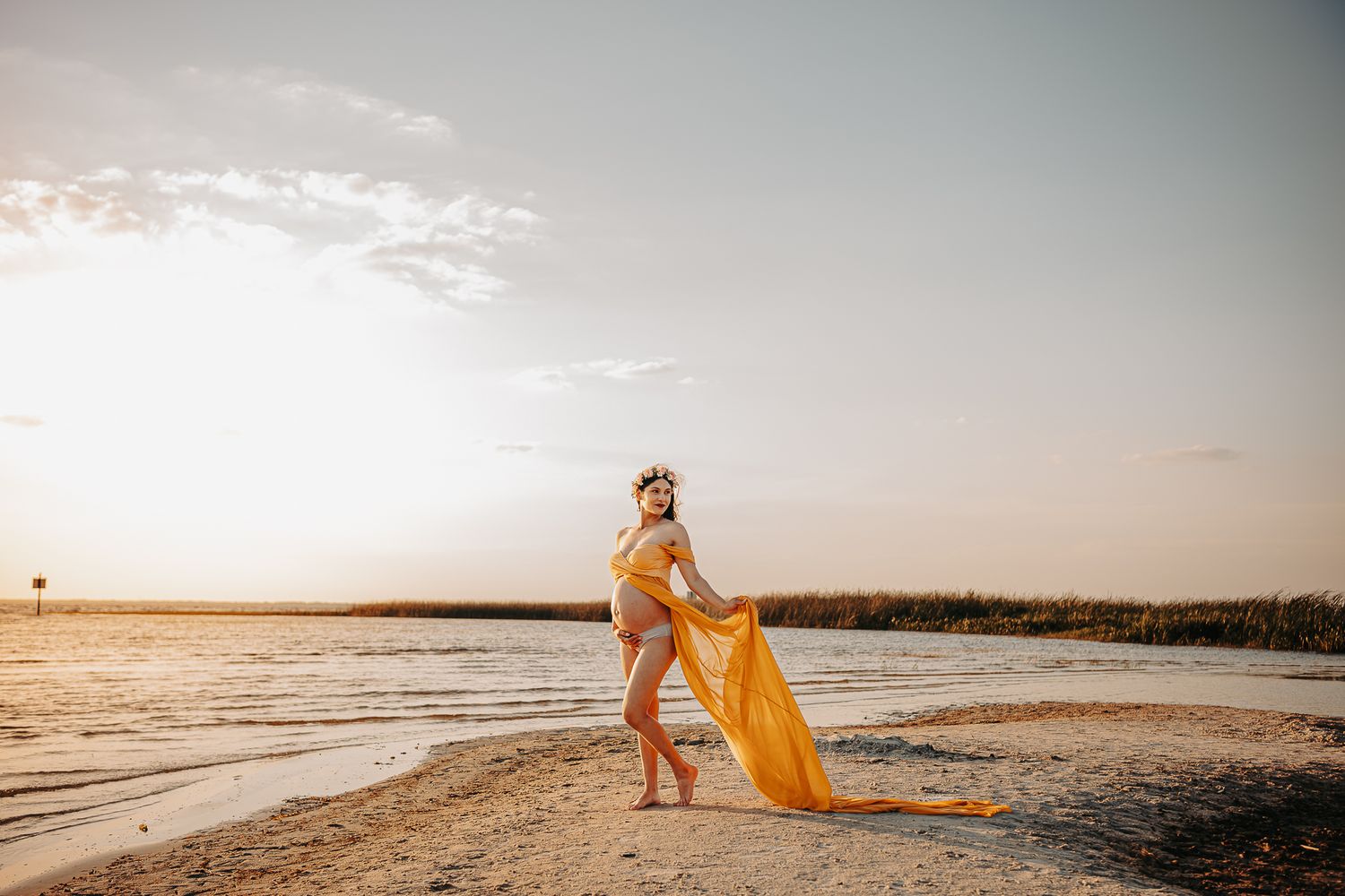 A woman in a flowing yellow dress stands on a beach holding a surfboard at sunset.
