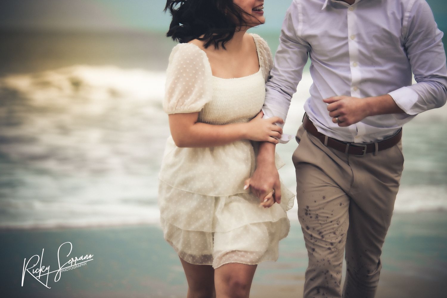 A couple strolls intimately along the shoreline wearing light colored clothing at dusk.