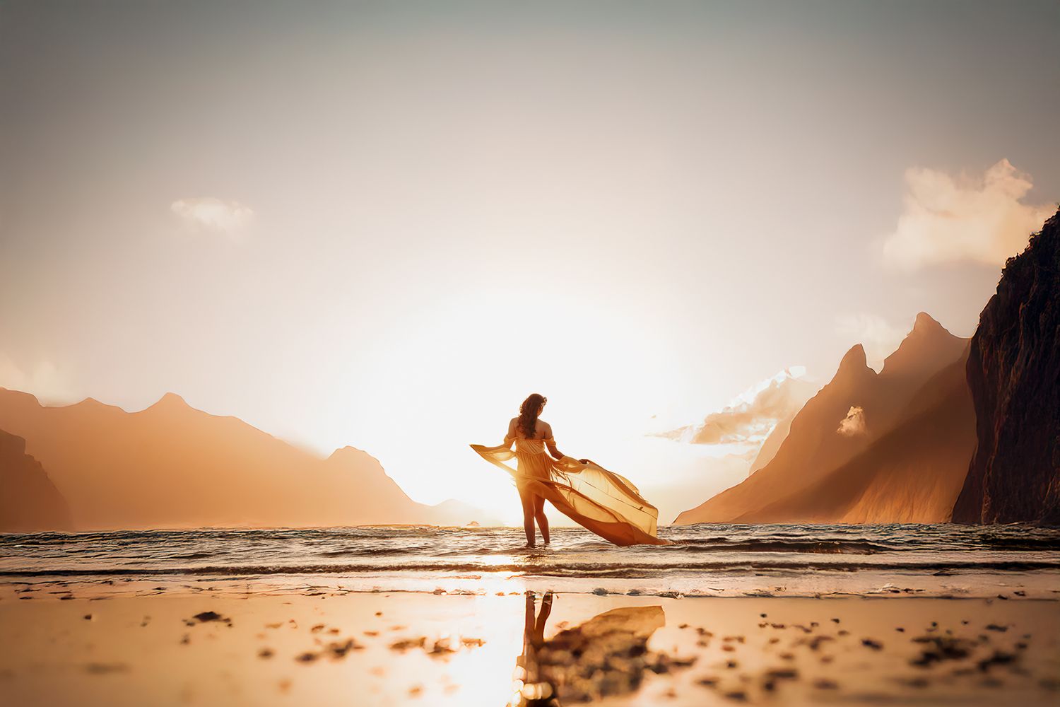 A silhouetted surfer holding their board on a beach at sunset with dramatic mountain peaks in the background.