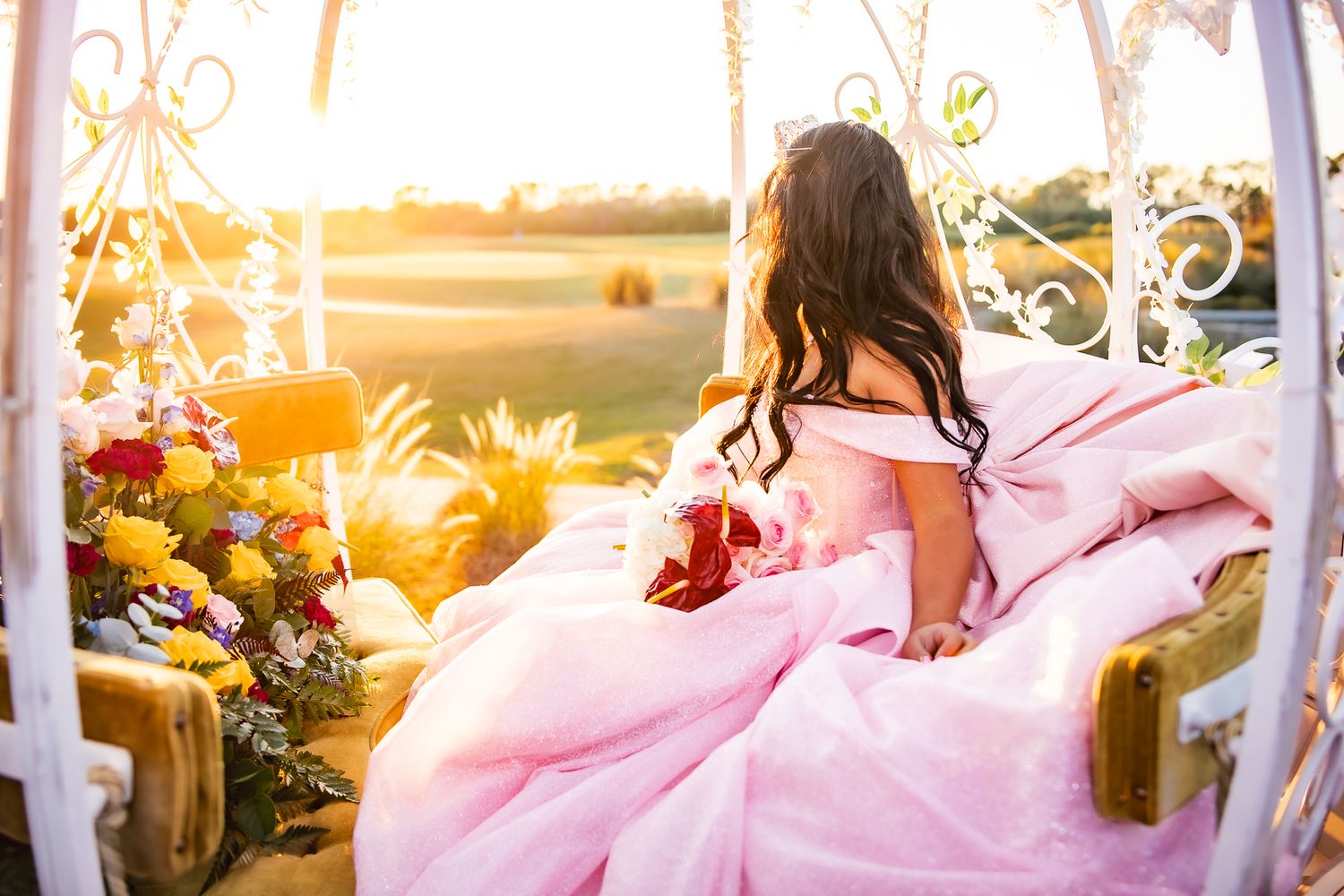 Person in flowing pink gown sits in vintage chair at sunset surrounded by flowers.