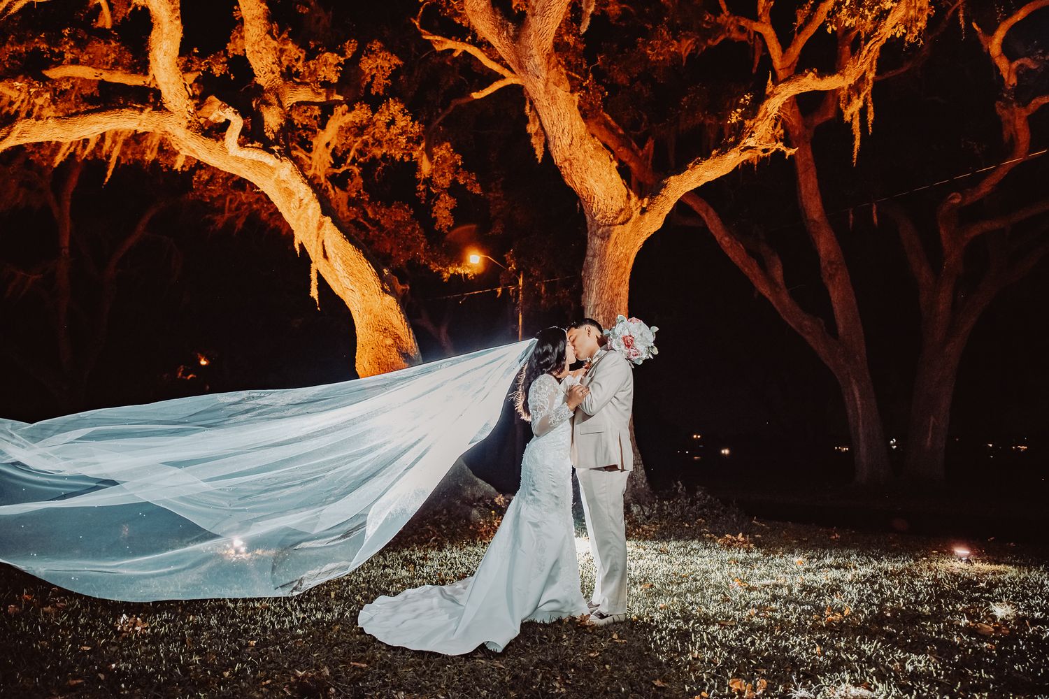 Long flowing white wedding veil floats dramatically against illuminated trees at night during romantic outdoor ceremony.