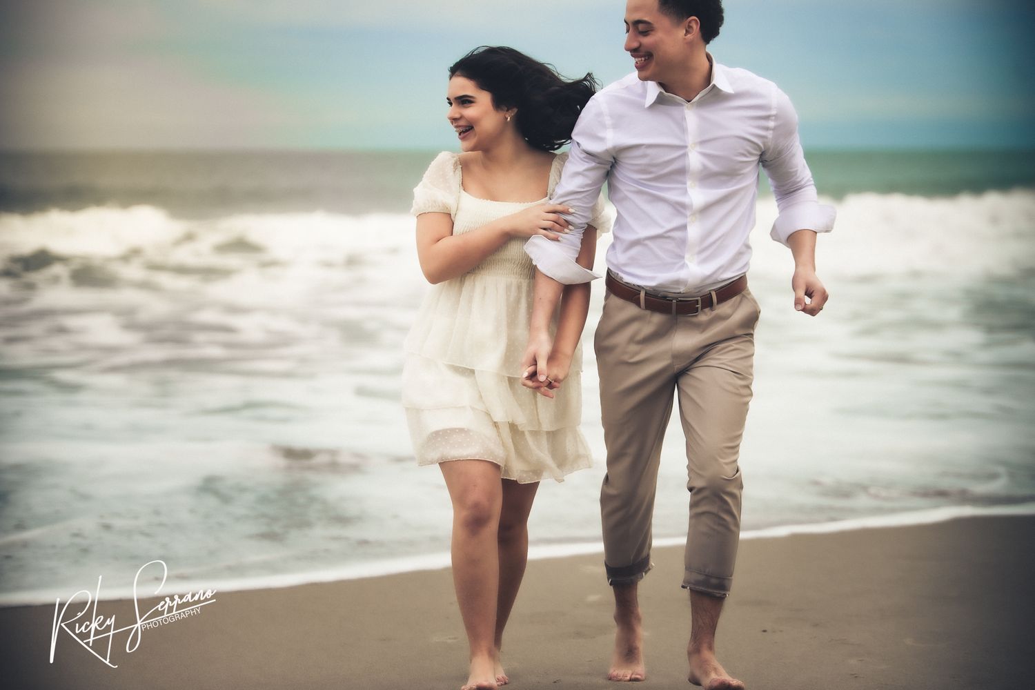 A romantic couple walks barefoot along a sandy beach at sunset, wearing casual attire.