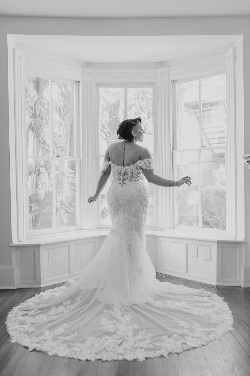 Black and white photograph of a bride in a lace mermaid wedding gown standing in a bay window with dramatic train.
