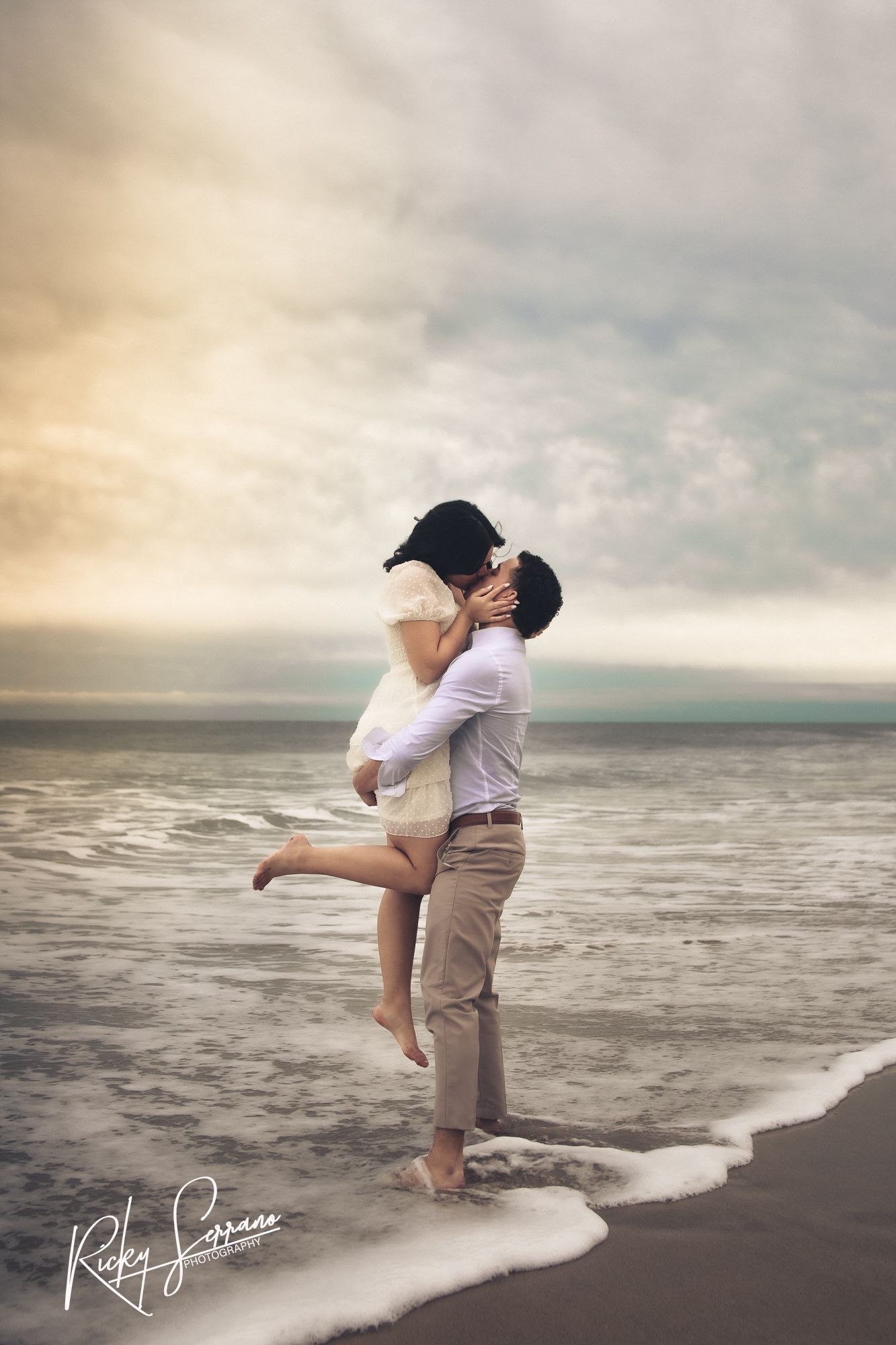 Romantic beach scene at sunset with a couple embracing on the shoreline as gentle waves lap at their feet.
