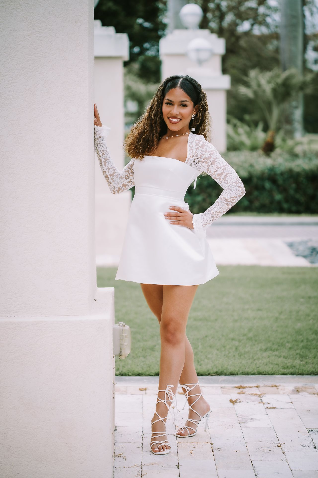 A woman in a short white lace-sleeved dress and strappy heels poses confidently against a white pillar.