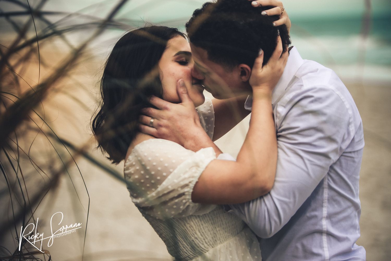 A romantic couple shares an intimate kiss outdoors, with the woman wearing a white lace dress and the man in a light shirt.