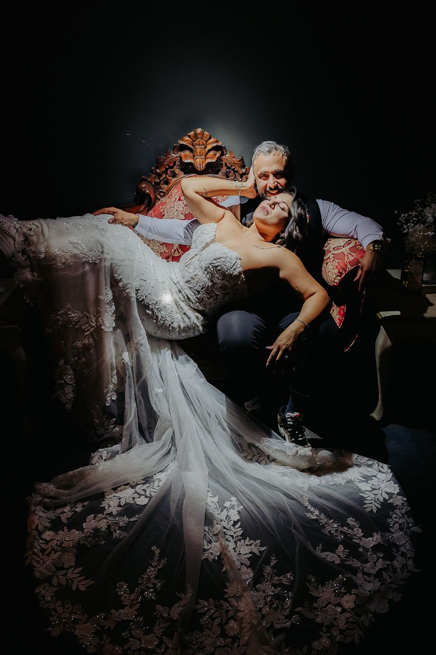 A couple shares a romantic pose on a vintage chaise lounge, with the bride's flowing lace gown cascading dramatically.