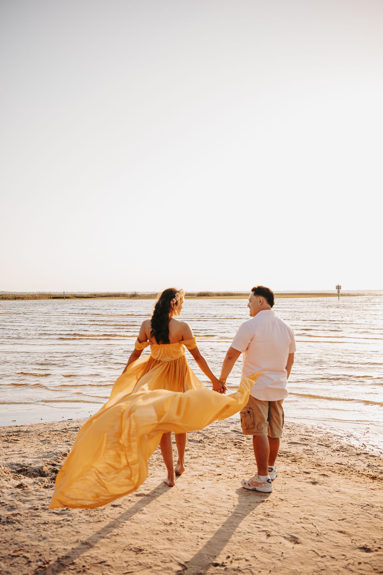 Couple holding hands walks on beach at sunset with flowing yellow dress catching the golden light.