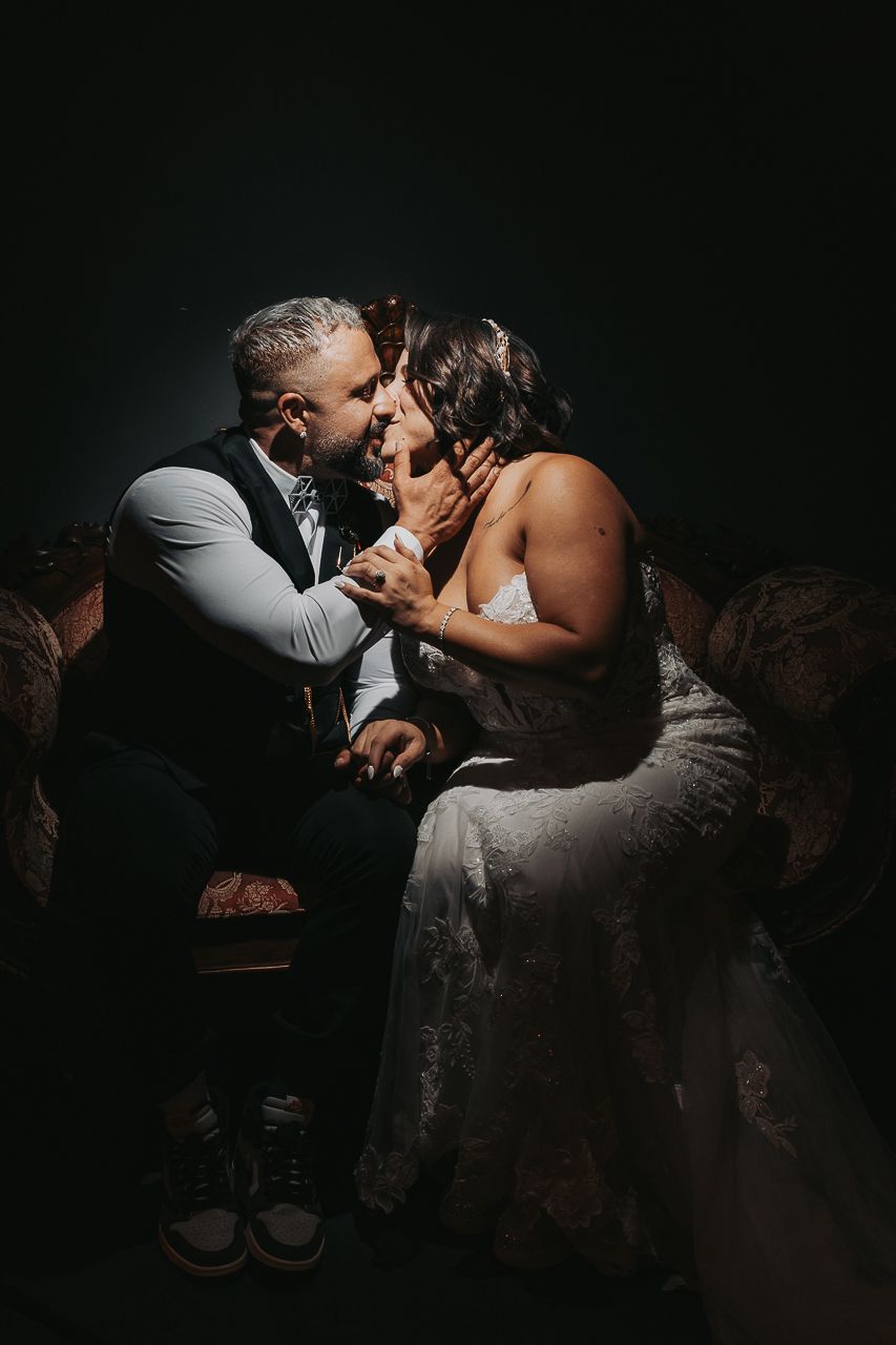 Romantic couple shares intimate moment in dark room, seated together on vintage sofa in dramatic moody lighting.