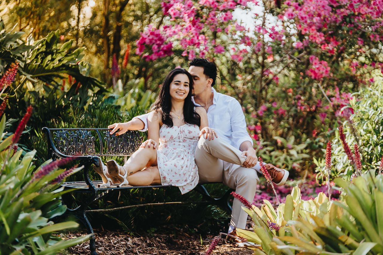 A romantic couple sits on a bench surrounded by vibrant pink bougainvillea flowers and tropical garden foliage.