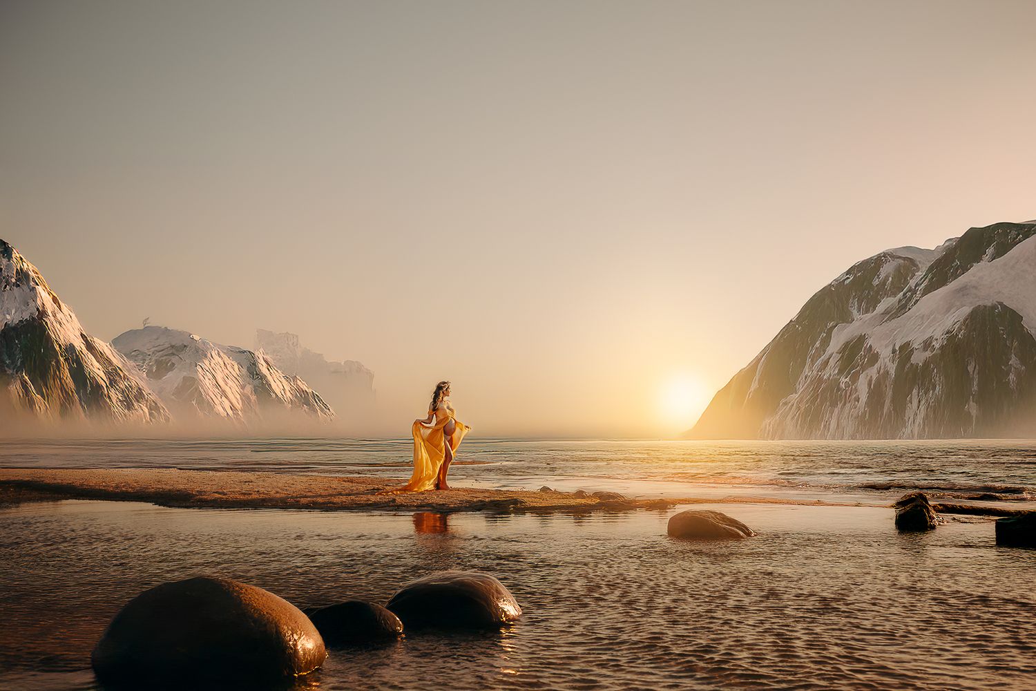 Person in yellow coat stands on rocky beach at sunset with snow-capped mountains and ocean in background.