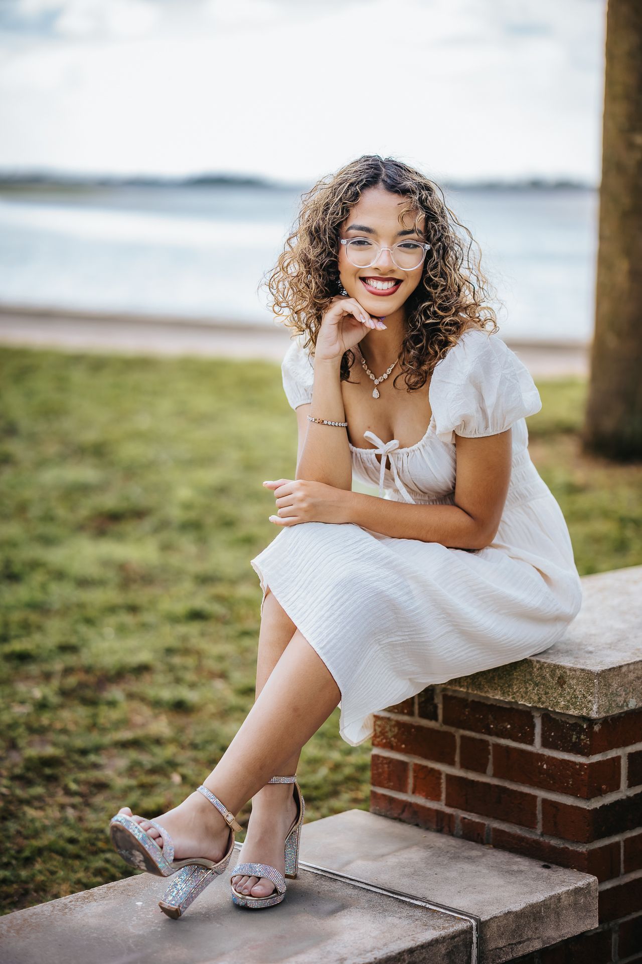 A woman in a white summer dress sits on a brick wall by the beach with natural curly hair.
