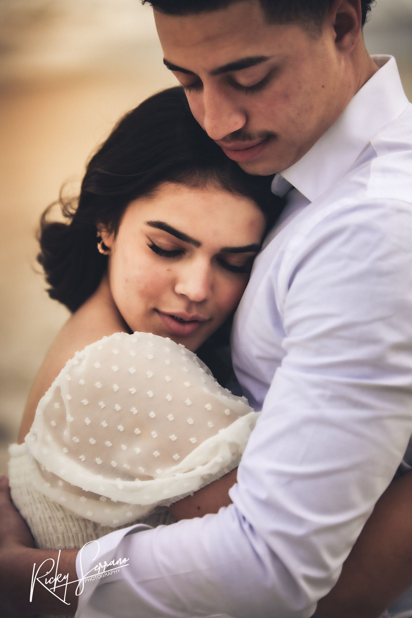 A romantic embrace between a couple in formal attire, with the person in a polka dot dress resting against their partner's chest.