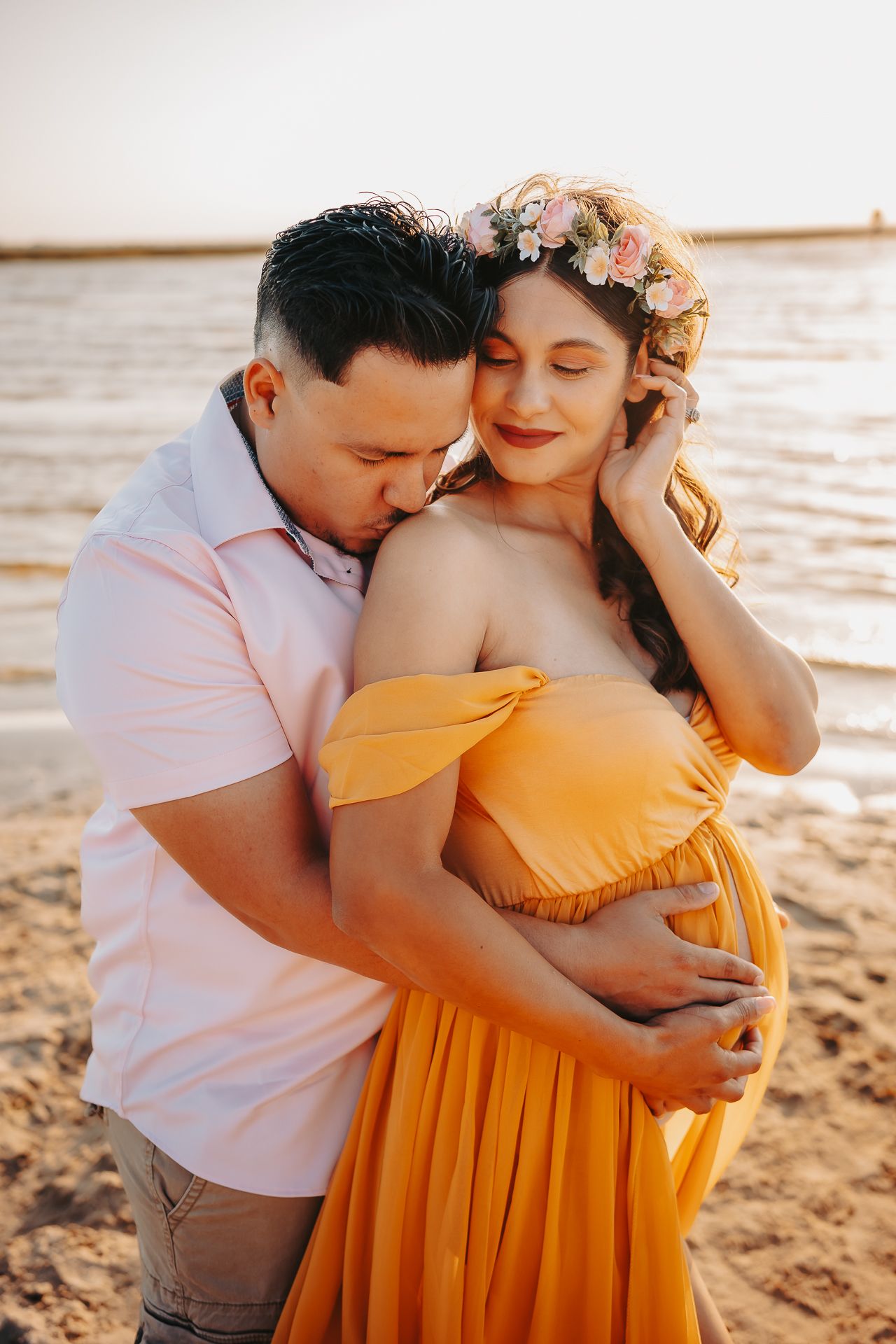 A tender maternity photoshoot moment at sunset on the beach, featuring a flowing yellow dress and floral crown.