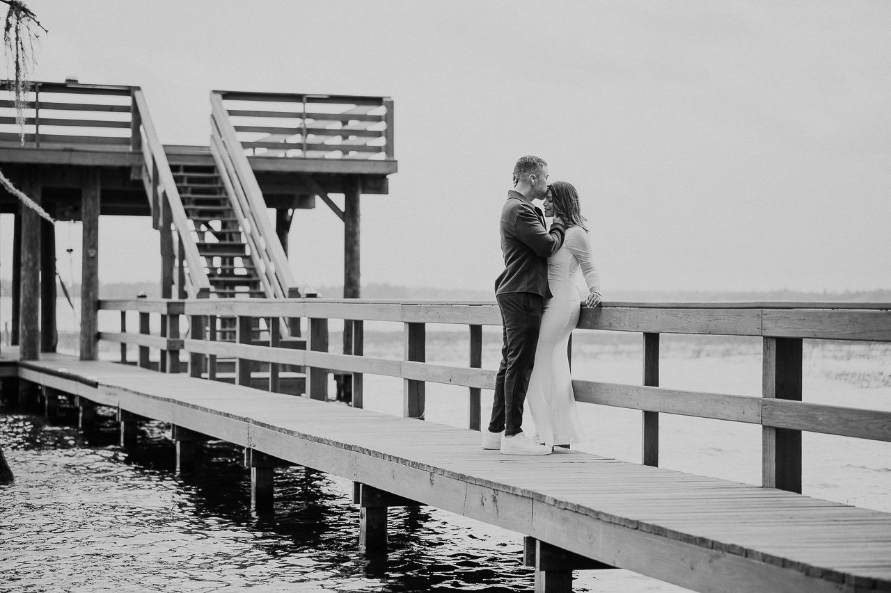 A couple embrace romantically on a wooden pier overlooking the water in a timeless black and white moment.