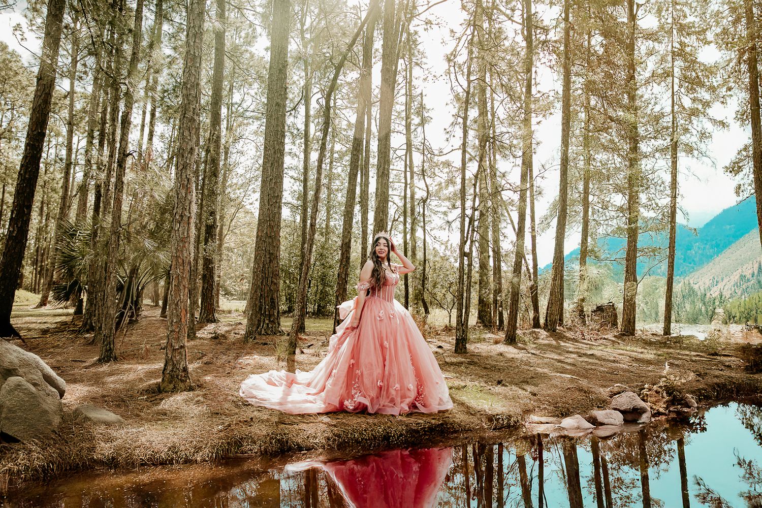 Person in pink ballgown poses reflectively by forest pond in mountain setting.