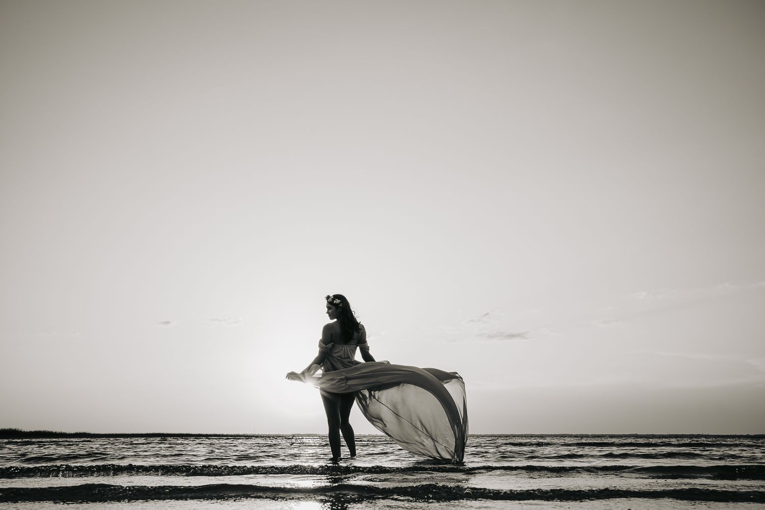 A silhouetted figure in a flowing dress dances gracefully on a beach at sunset in black and white photography.
