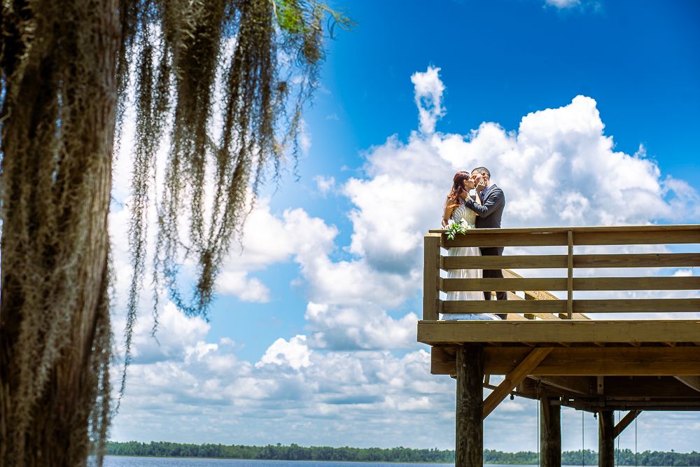A person enjoys a peaceful moment on a wooden lakeside deck under Spanish moss and puffy clouds in Florida.
