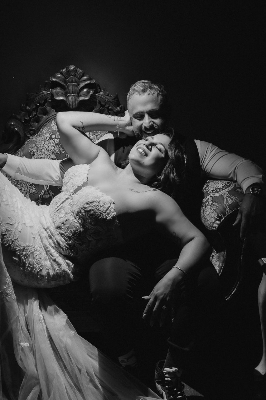 Black and white wedding portrait showing a romantic moment with the bride in a lace dress reclining on an ornate sofa.