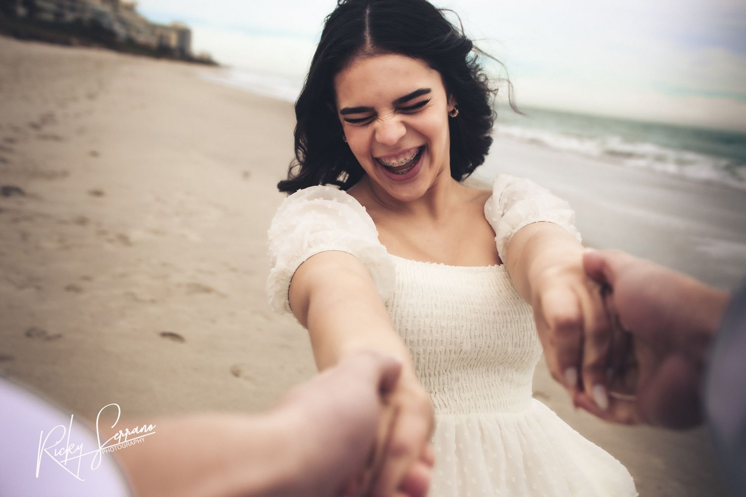 Person in white dress laughs joyfully while spinning on a cloudy beach with waves in the background.