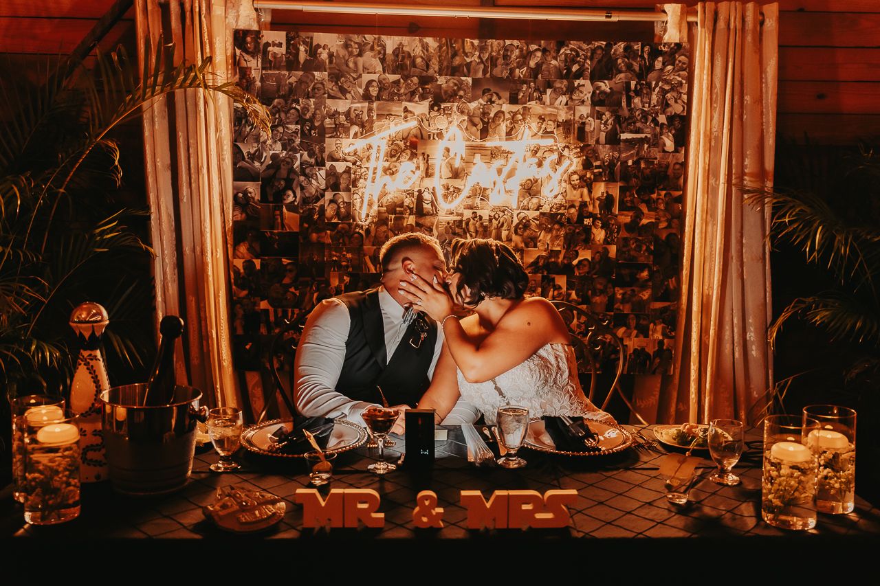 A romantic wedding couple sits on floor with glowing lanterns and floral backdrop at nighttime reception.