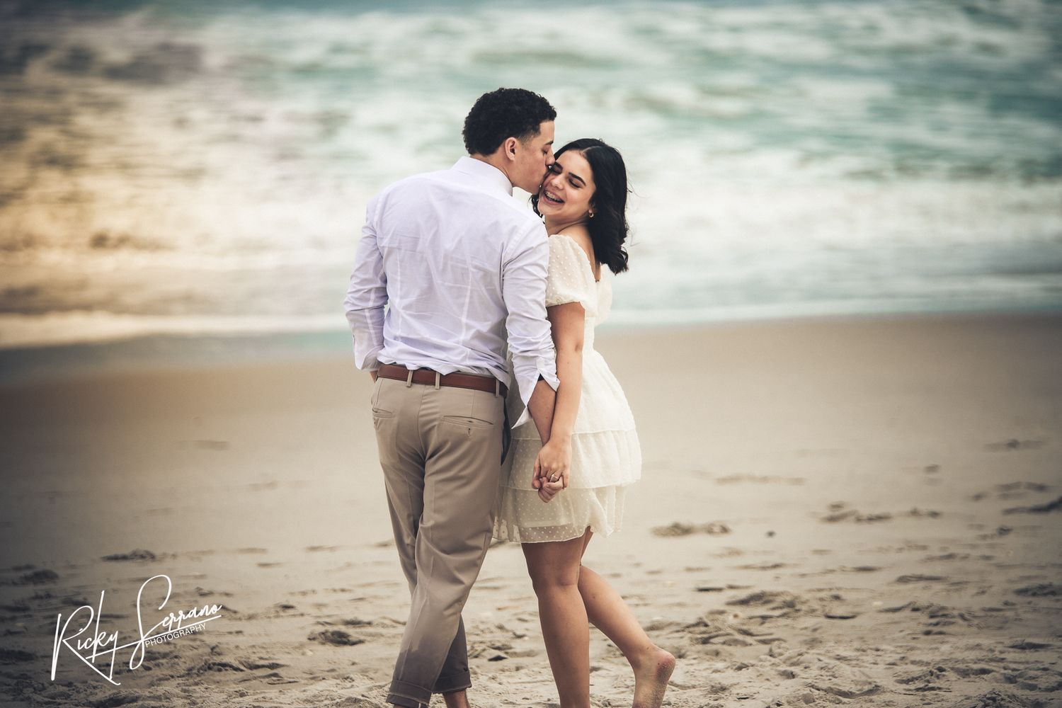 A couple in light colored clothing share an intimate moment while walking on a beach at sunset.