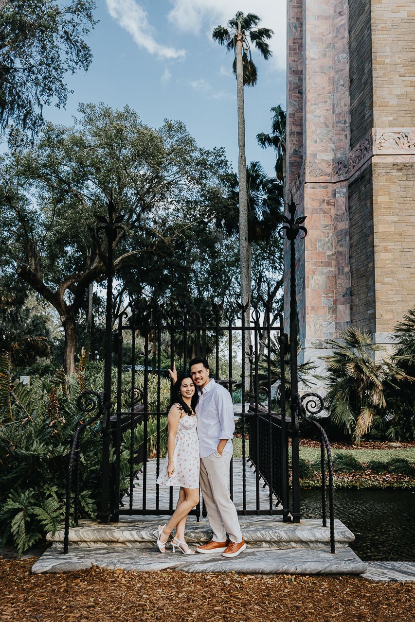 A couple embraces at an ornate iron gate entrance with a tall palm tree and lush greenery in the background.
