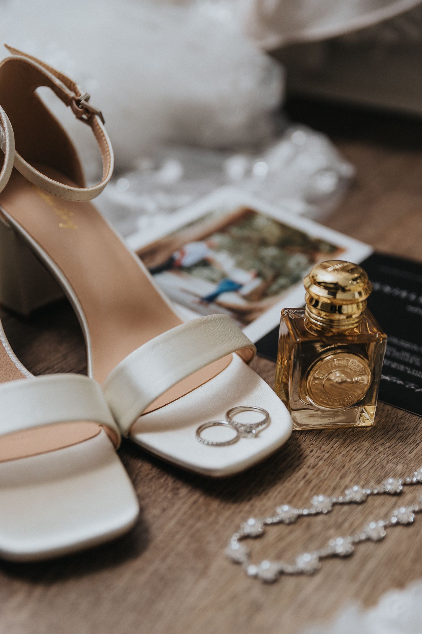 Wedding accessories including white heels, rings and perfume bottle arranged on wooden surface.