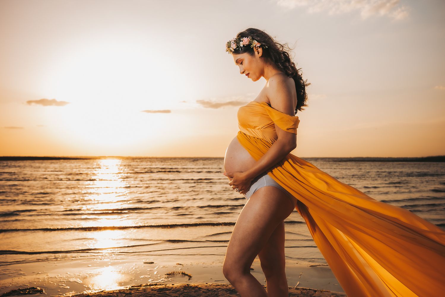 Silhouetted pregnant figure in flowing yellow dress stands at beach during golden sunset.