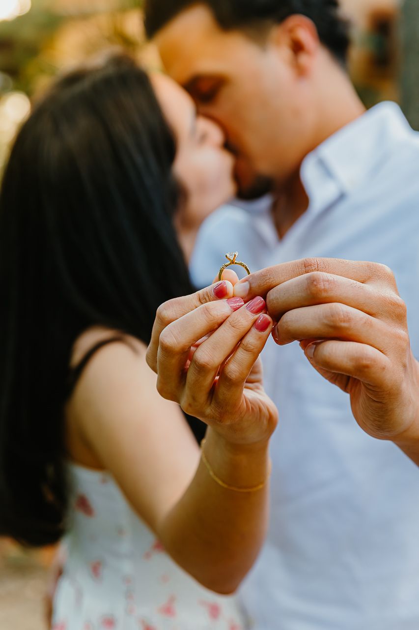 Romantic proposal moment with hands holding an engagement ring while a couple shares an intimate kiss in soft lighting.