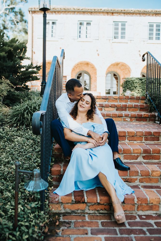 A couple sits together on brick steps outside a Mediterranean-style building with arched windows and blue railings.